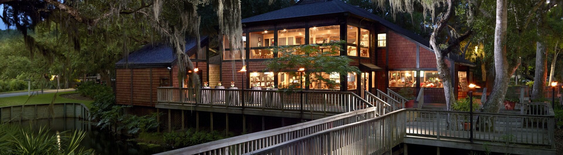 A large wooden lodge with many windows, surrounded by trees and greenery, is lit warmly at dusk. A wooden walkway leads to the entrance over water with lush plants and moss-draped branches.