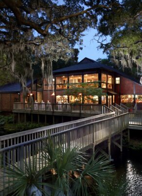 A large wooden lodge with many windows, surrounded by trees and greenery, is lit warmly at dusk. A wooden walkway leads to the entrance over water with lush plants and moss-draped branches.