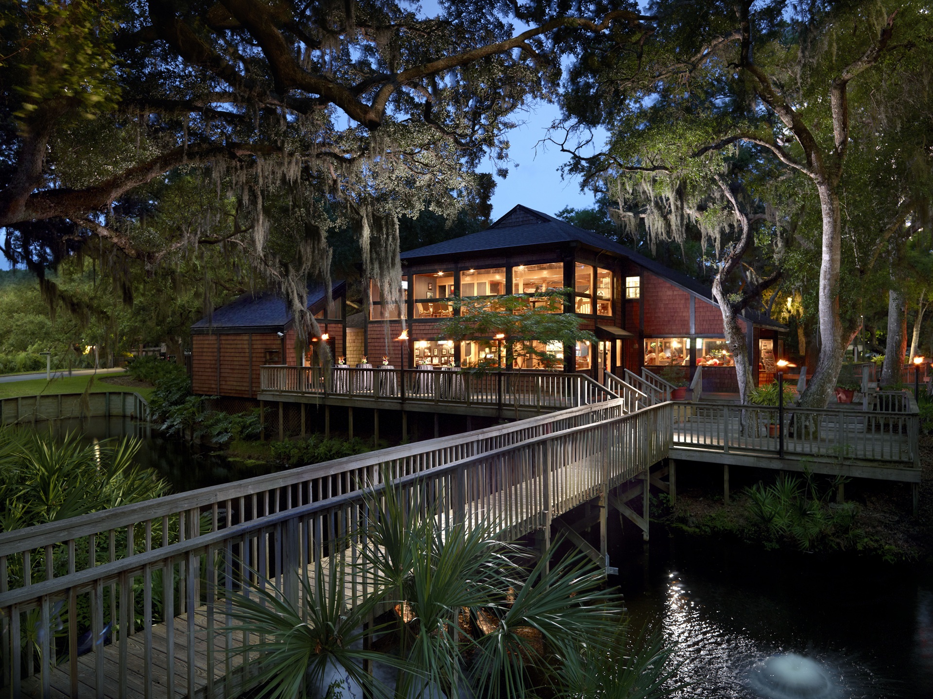 A large wooden lodge with many windows, surrounded by trees and greenery, is lit warmly at dusk. A wooden walkway leads to the entrance over water with lush plants and moss-draped branches.