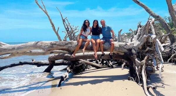 Three people sit on a large driftwood tree on a sandy beach, smiling at the camera. The sky is clear and blue, with the ocean and more driftwood in the background.