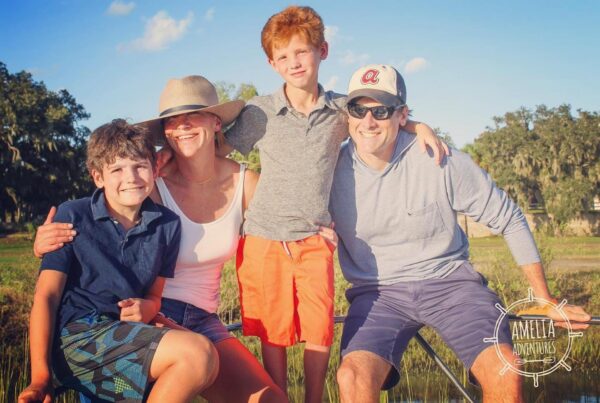 A smiling family of four poses outdoors on a sunny day, with trees and blue sky in the background. The parents wear casual summer clothes and hats, while the two boys stand and sit close, all looking happy. Amelia Adventures logo is visible.