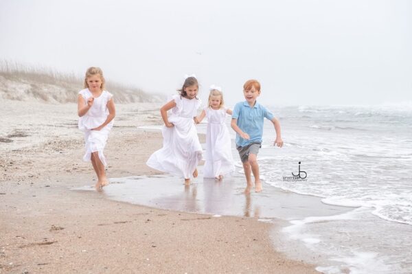 Four young children, three girls in white dresses and one boy in a blue shirt and shorts, run and laugh along a sandy, misty beach with gentle waves in the background.