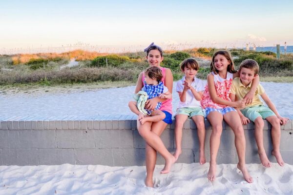 Five children sit on a low brick wall at a beach, with sand in the foreground and grassy dunes behind them. They smile and look relaxed, with the sunset sky giving a warm, gentle light.