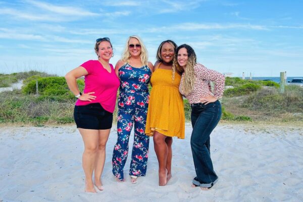 Four women smile and pose together on a sandy beach, with greenery and a blue sky in the background. They wear colorful summer outfits and appear happy and relaxed.