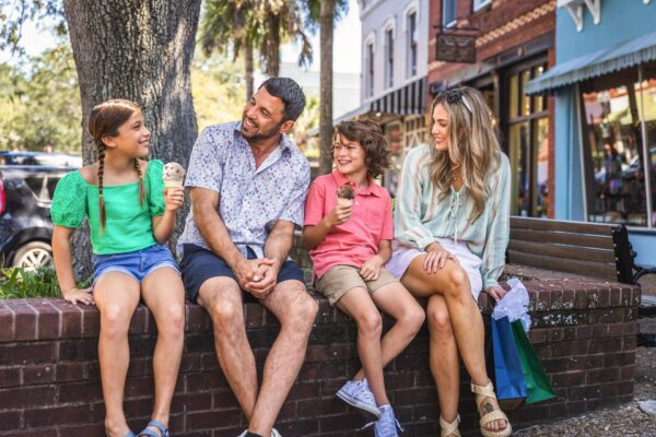 A family of four sits on a brick wall outdoors, smiling and eating ice cream together. Shops and trees are visible in the background, and the mother holds shopping bags. It appears to be a sunny day.