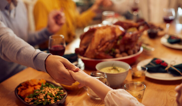 Family members hold hands around a dinner table set with Thanksgiving food, including roast turkey, sweet potatoes, green beans, and wine glasses, creating a warm, festive atmosphere.