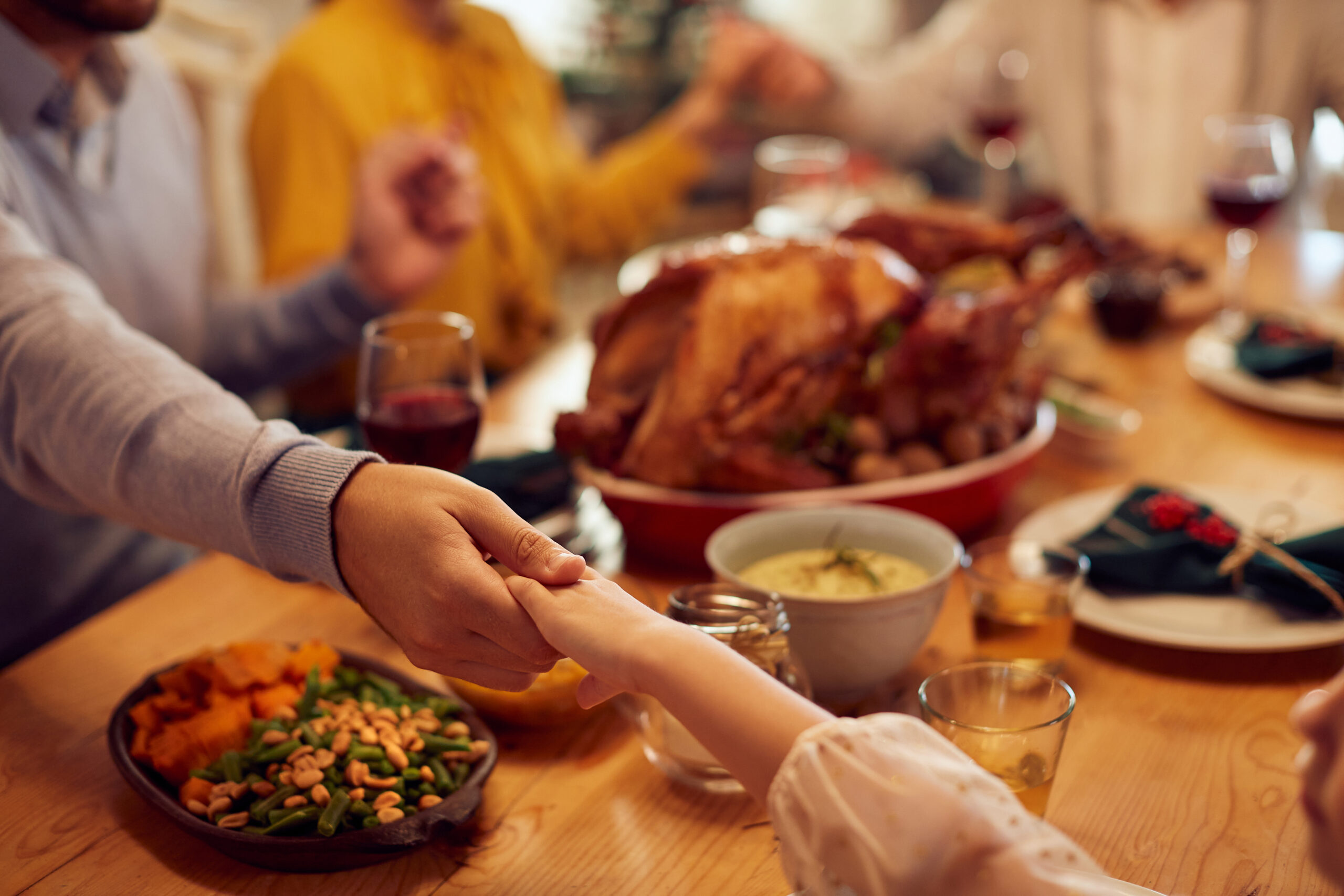 Family members hold hands around a dinner table set with Thanksgiving food, including roast turkey, sweet potatoes, green beans, and wine glasses, creating a warm, festive atmosphere.