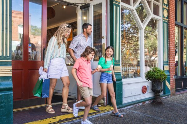 A smiling family of four, two adults and two children, walk out of a store with shopping bags on a sunny day in a charming, historic downtown area.
