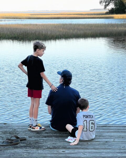 Two children and an adult sit on a wooden dock by calm water, with tall grass and trees in the distance. One child stands beside the adult, while the other sits close, touching the adults arm. Sandals lie nearby.