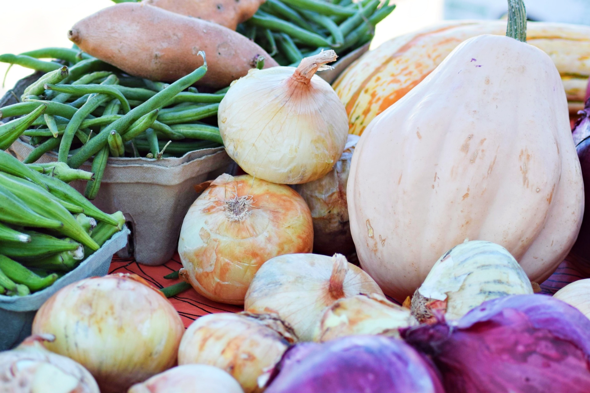 A close-up of assorted fresh vegetables, including onions, green beans, sweet potatoes, red onions, and a pale butternut squash, displayed together on a table.