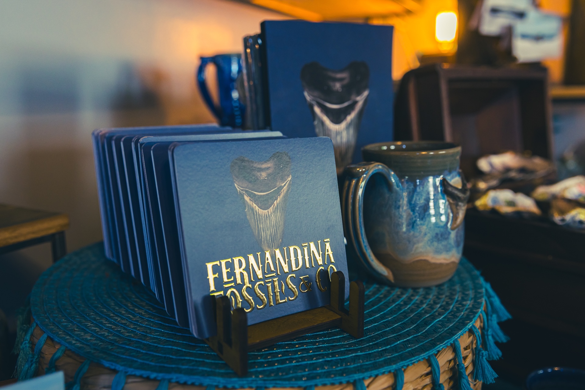 A display of blue coasters featuring a fossilized shark tooth illustration, with a sign reading Fernandina Fossils & Co, next to a ceramic mug on a woven blue tablecloth in a warmly lit shop.