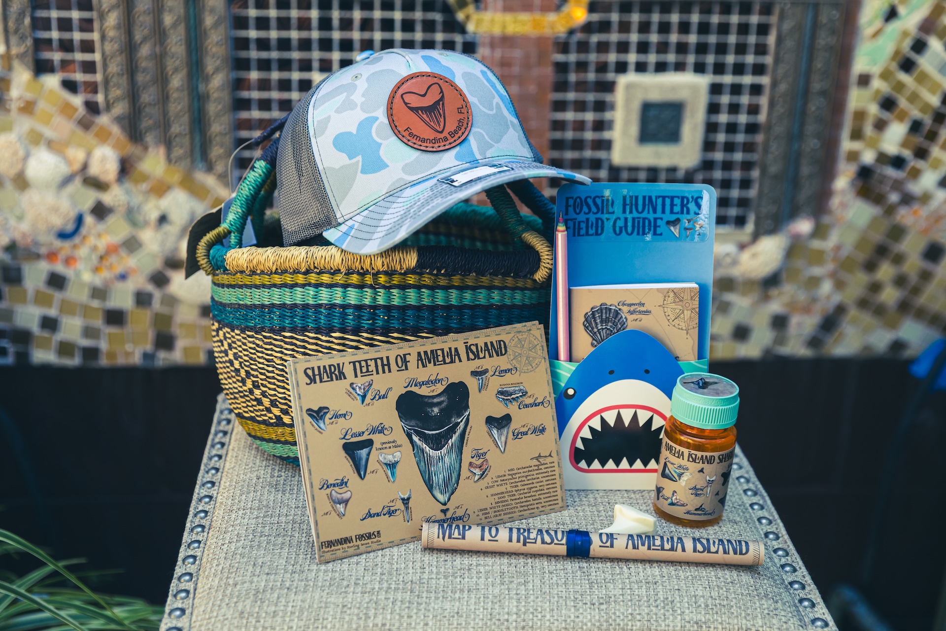 A collection of shark-themed items, including a cap, a woven basket, postcards, a shark illustration, a field guide, a small bottle, and a rolled map, displayed on a patterned cloth against a mosaic wall.