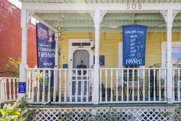 A yellow house with white trim and a porch displays banners reading Fernandina Fossils Museum and Now Open! Fossils! A blue handicap accessible sign is attached to the railing near the entrance.