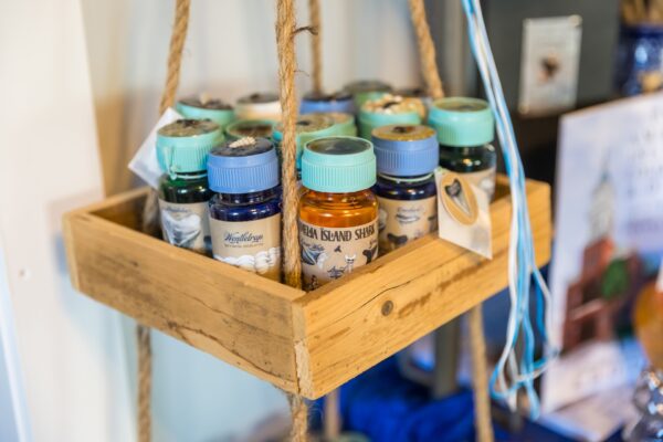 A wooden hanging shelf holds several small glass bottles with blue and green lids, labeled with beach-themed designs, alongside seashell souvenirs.
