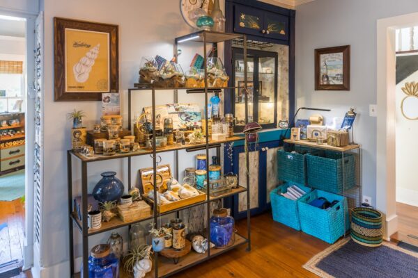 A cozy room with shelves displaying assorted decorative items, jars, and sea-themed trinkets. Blue baskets with towels are on the floor, and a blue cabinet stands against a light wall with framed art.
