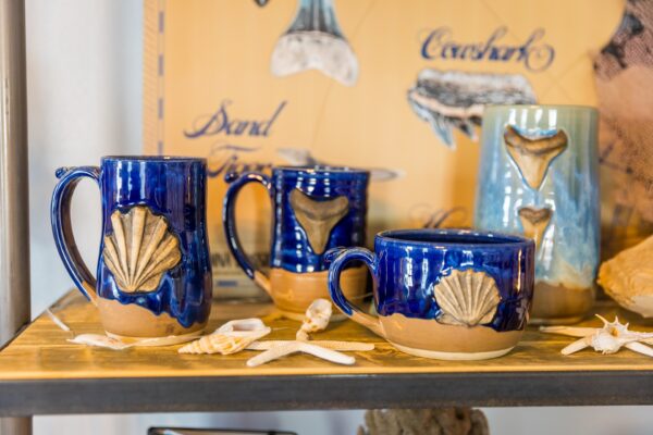Blue ceramic mugs and a vase with beige seashell decorations are displayed on a wooden shelf, surrounded by assorted seashells and starfish, with a nautical-themed background.
