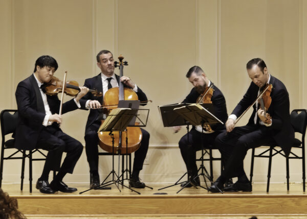 Four musicians in black suits sit onstage playing string instruments—two violins, a cello, and a viola. They read sheet music on stands and appear focused, performing in a formal concert setting.