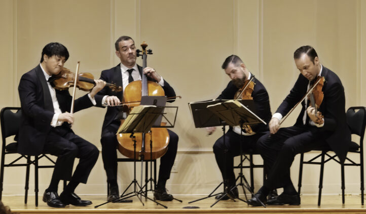 Four musicians in black suits sit onstage playing string instruments—two violins, a cello, and a viola. They read sheet music on stands and appear focused, performing in a formal concert setting.