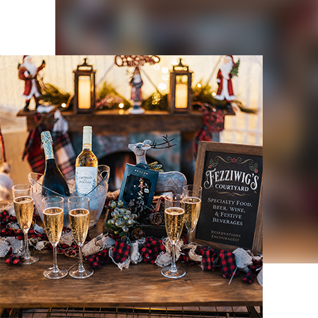 A festive display with sparkling wine, glasses, a Fezziwigs Courtyard sign, holiday decor, lanterns, and Santa figurines on a wooden table, creating a cozy Christmas atmosphere.