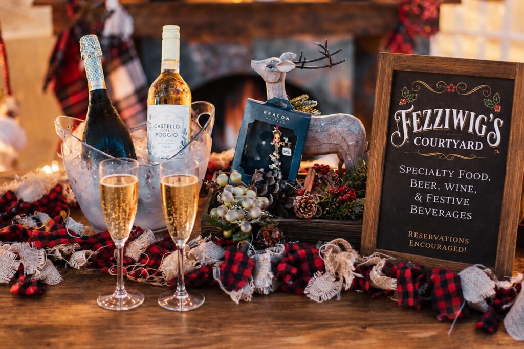 Two glasses of sparkling wine next to an ice bucket with wine bottles, a decorative reindeer, and a chalkboard sign for Fezziwigs Courtyard listing festive beverages.