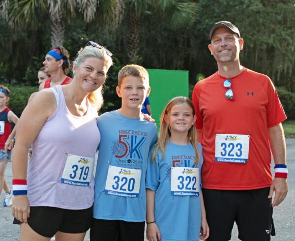 A smiling family of four poses outdoors at a race event, all wearing numbered bibs. The parents wear athletic clothes and wristbands; the children wear matching blue Firecracker 5K shirts. Trees are visible in the background.