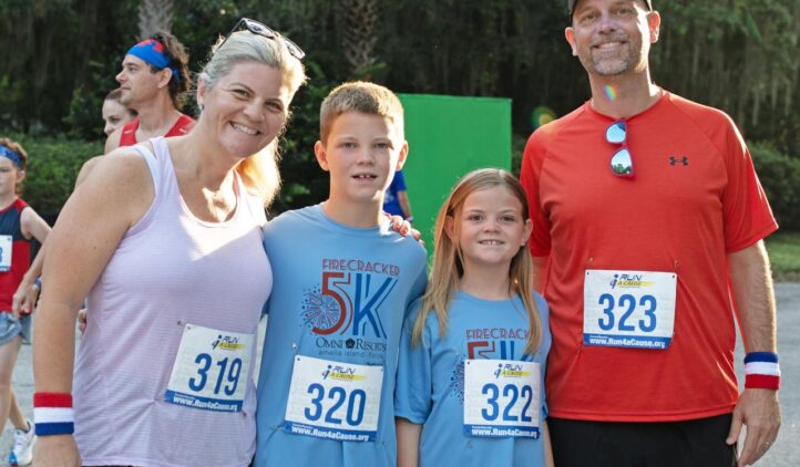 A smiling family of four poses outdoors at a race event, all wearing numbered bibs. The parents wear athletic clothes and wristbands; the children wear matching blue Firecracker 5K shirts. Trees are visible in the background.