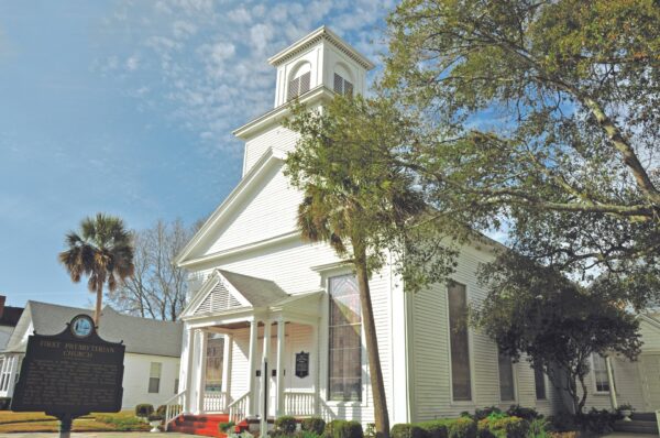 A white wooden church with tall windows, a steeple, and a front porch sits on a sunny day, surrounded by trees. A historical marker stands on the lawn near the entrance.