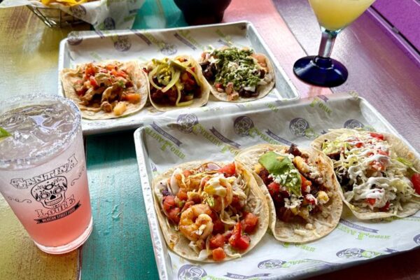 Three trays with assorted tacos filled with shrimp, beef, chicken, and veggies sit on a colorful table. A pink drink in a Tantina Louie’s cup and a margarita in a blue-rimmed glass are nearby.