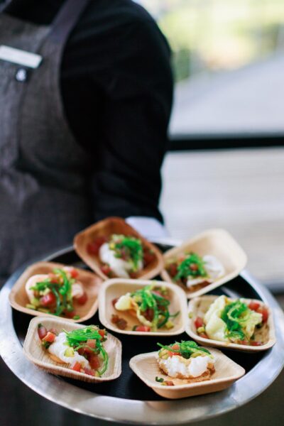 A server in a black shirt and gray apron carries a round tray with several small wooden dishes, each holding a colorful appetizer garnished with green vegetables and diced tomatoes.