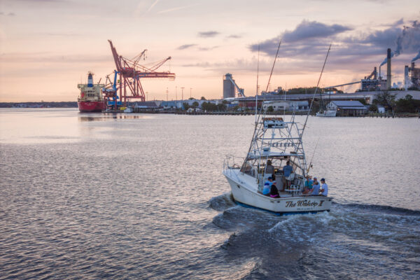 A small fishing boat named The Wahoo II carries several people across a harbor at sunset, with industrial cranes, a large cargo ship, and factory buildings visible in the background.