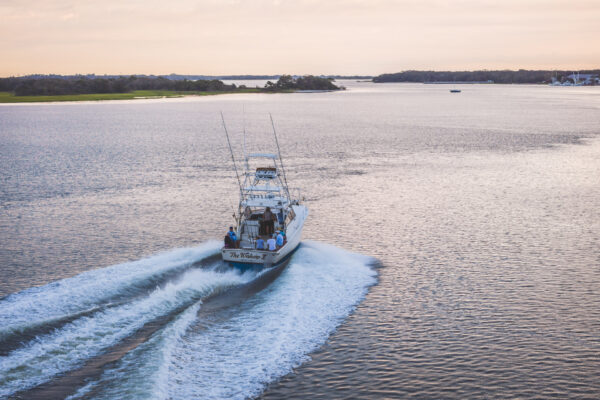 A white fishing boat moves away quickly, leaving a wake on calm water near a shoreline with trees under a pastel sky at sunset.