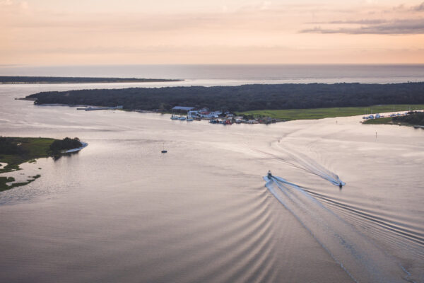 Aerial view of two boats creating wakes on a wide river at sunset, with a small island and buildings in the distance, surrounded by greenery and calm water under a pastel sky.