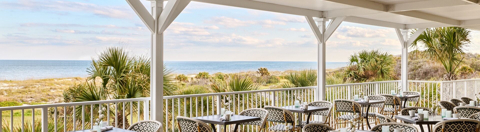 Covered outdoor restaurant patio with empty tables and chairs, overlooking palm trees, sand dunes, and the ocean under a partly cloudy sky.