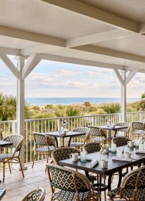 Covered outdoor restaurant patio with empty tables and chairs, overlooking palm trees, sand dunes, and the ocean under a partly cloudy sky.