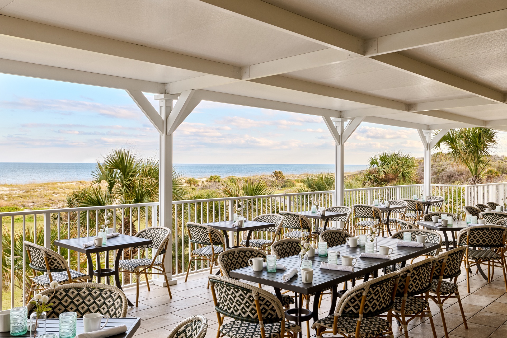 Covered outdoor restaurant patio with empty tables and chairs, overlooking palm trees, sand dunes, and the ocean under a partly cloudy sky.
