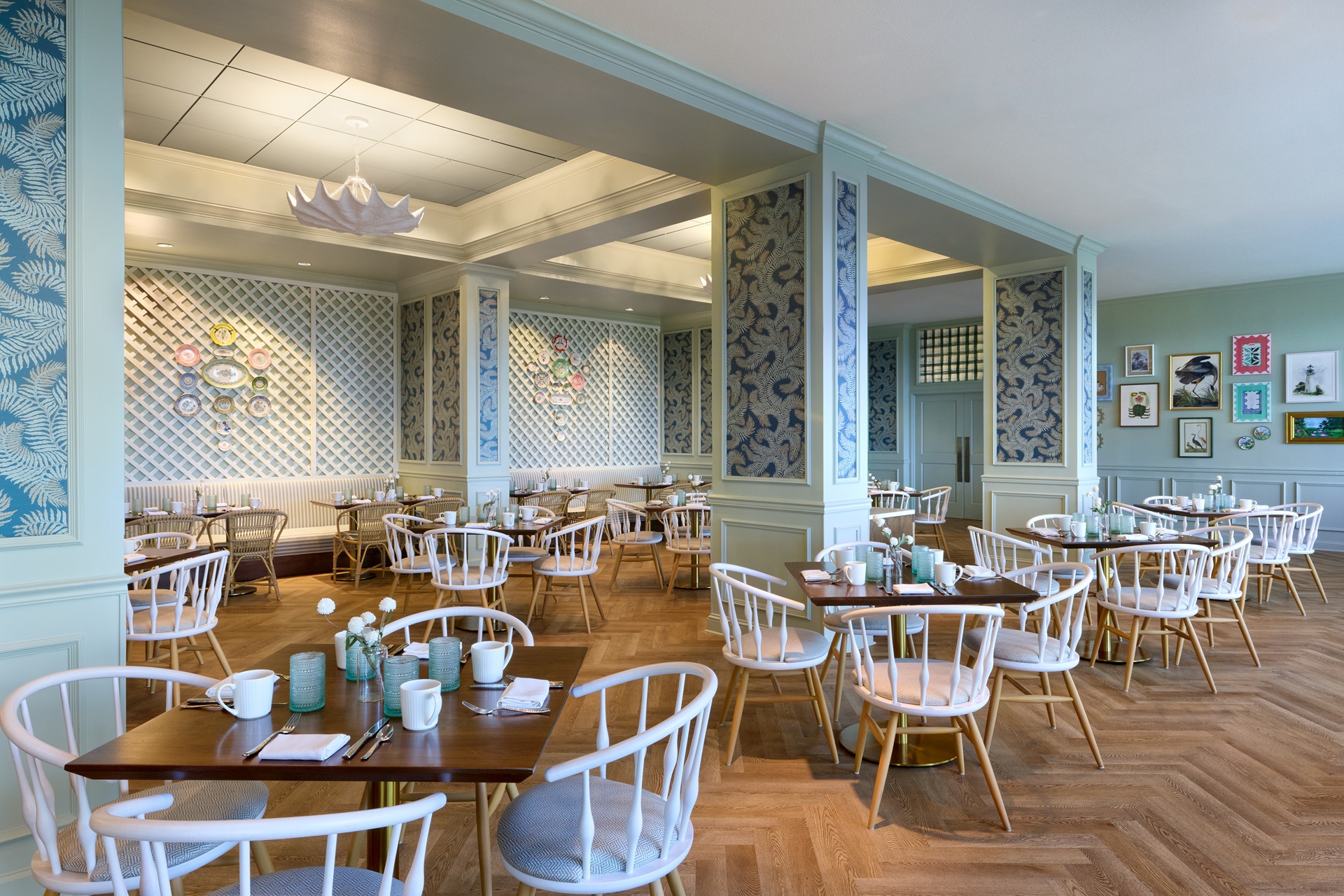 A bright, stylish cafe with wooden floors, light blue walls, white chairs, and wooden tables set for dining. Decorative plates and framed artwork adorn the walls. The atmosphere is cozy and inviting.