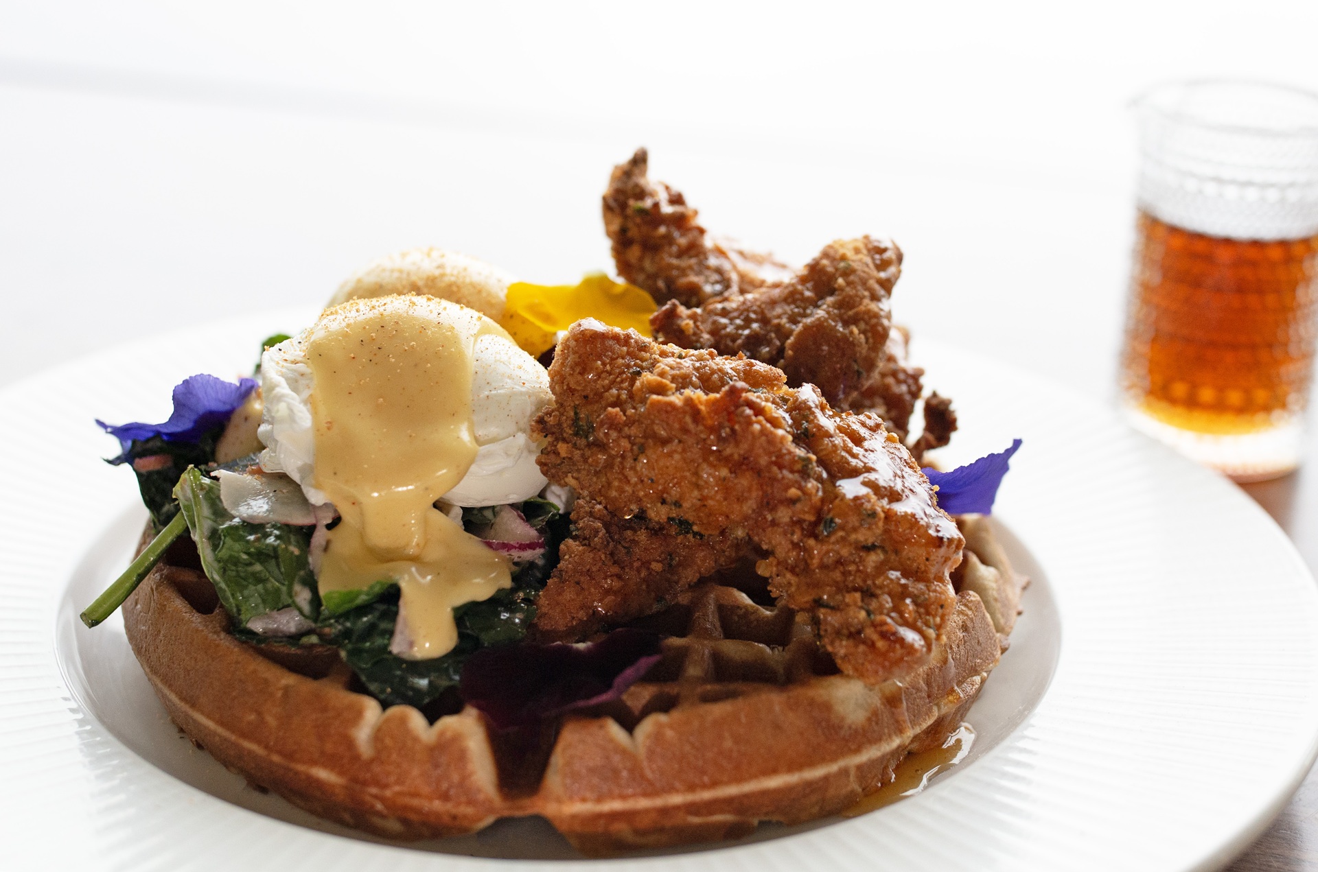 A plate with a waffle topped with fried chicken, poached egg, hollandaise sauce, sautéed greens, and edible flowers. A small glass of syrup is in the background.