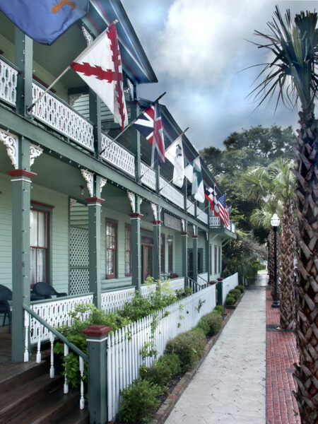 A historic green building with white trim and decorative railings displays several flags along a porch. Palm trees and a brick sidewalk line the street under a cloudy sky.