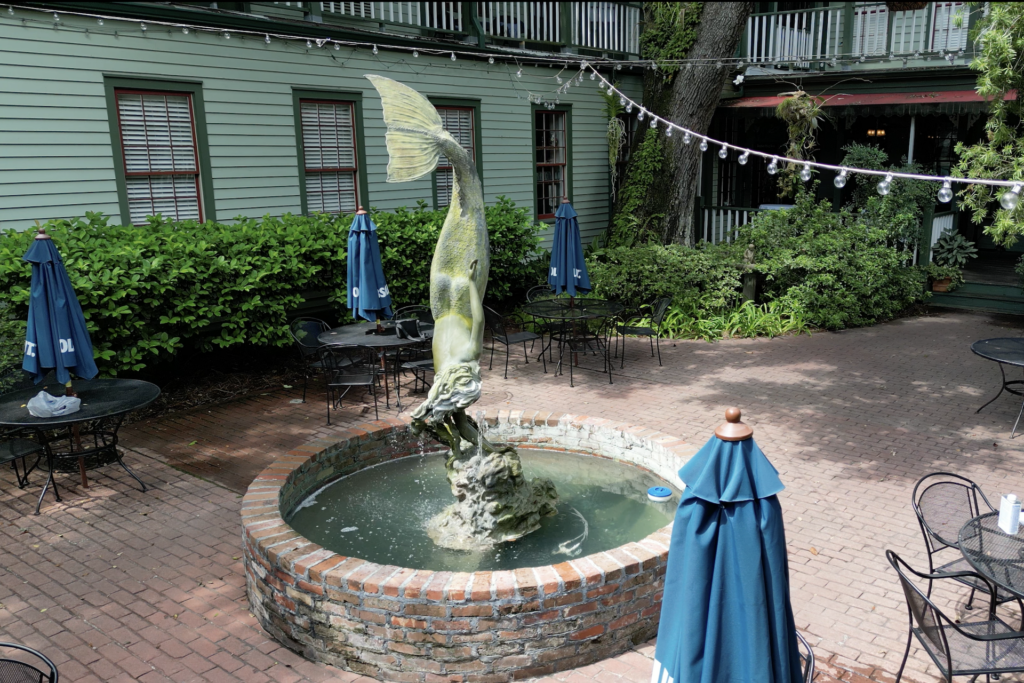 A small brick fountain with a large fish statue in the center of a patio, surrounded by tables, chairs, blue umbrellas, and greenery near a green building.