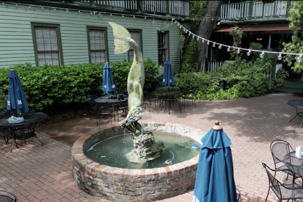 A small brick fountain with a large fish statue in the center of a patio, surrounded by tables, chairs, blue umbrellas, and greenery near a green building.