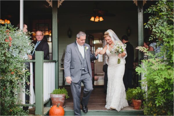 A bride in a white dress and veil holds a bouquet and smiles as an older man, possibly her father, helps her down the steps of a porch. Guests watch in the background, and a pumpkin sits by the stairs.