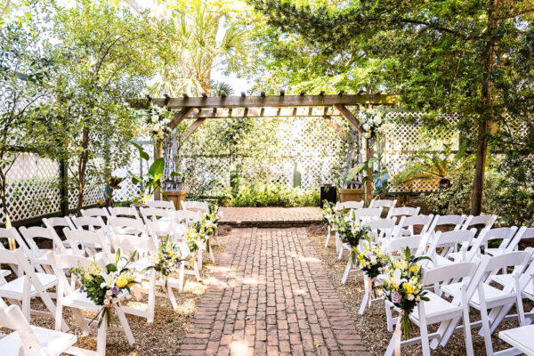 An outdoor wedding ceremony setup featuring rows of white chairs with floral arrangements, facing a wooden pergola adorned with flowers, surrounded by lush greenery and lattice fencing.