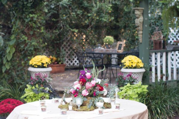 A round table with a floral centerpiece and candles sits in a garden, surrounded by lush greenery and colorful potted flowers. In the background, there’s a small patio table and chairs with decorative lights.