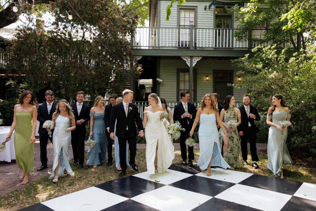 A wedding party poses outdoors in front of a green house, with bridesmaids in pastel dresses and groomsmen in dark suits. The bride and groom stand in the center, holding hands, on a large black-and-white checkered floor.