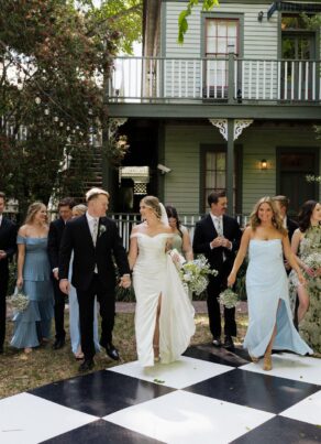 A wedding party poses outdoors in front of a green house, with bridesmaids in pastel dresses and groomsmen in dark suits. The bride and groom stand in the center, holding hands, on a large black-and-white checkered floor.