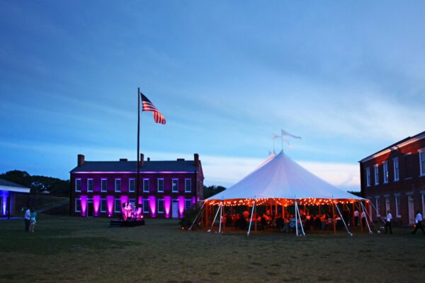 A large white tent with string lights glows at dusk between brick buildings. An American flag on a tall pole stands nearby, and purple lights illuminate the buildings. People gather under the tent on the grassy lawn.