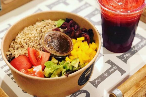 A bowl with quinoa, diced tomatoes, avocado, yellow bell peppers, black olives, and salad greens, served with a small container of dressing. A red juice drink in a plastic cup is next to the bowl.