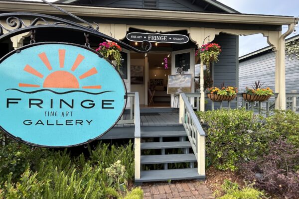 A bright turquoise sign for Fringe Fine Art Gallery hangs by the entrance of a gray house with white trim, surrounded by plants and hanging flower baskets. An OPEN sign is visible inside the doorway.