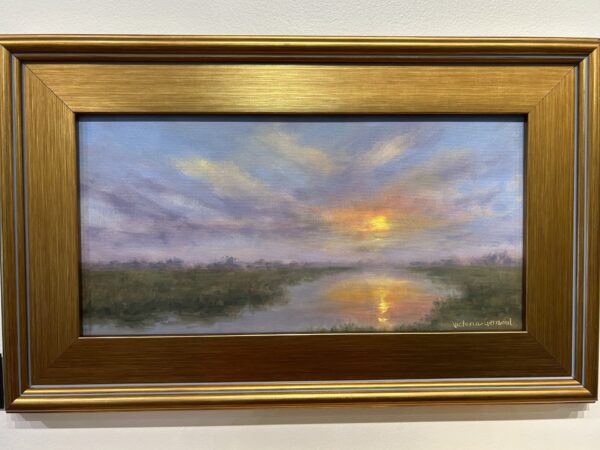 A framed painting of a serene landscape at sunset, with the sun low on the horizon, colorful clouds in the sky, and its reflection shining on a calm body of water surrounded by greenery.