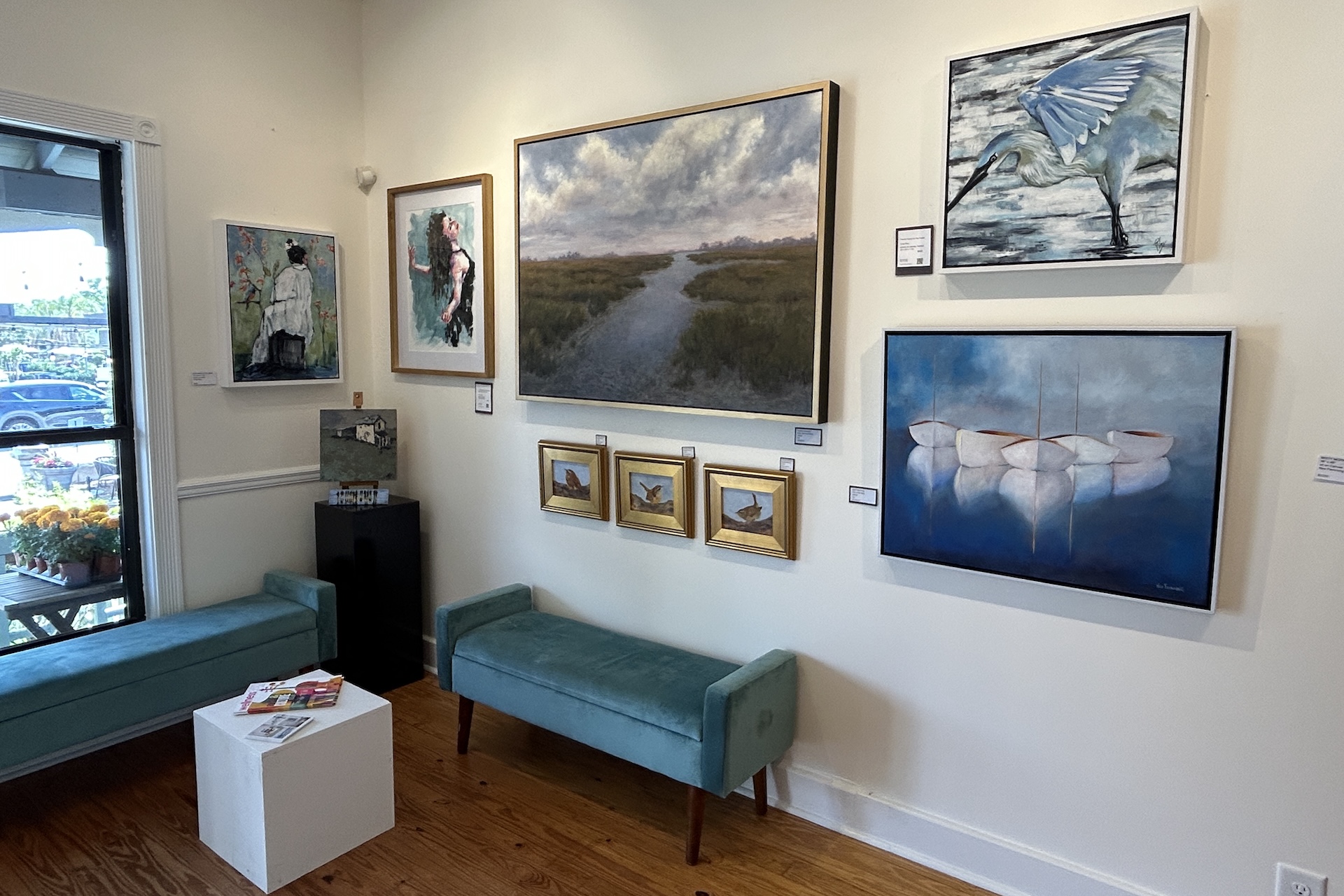 Art gallery interior with teal benches, a small white table, and various framed paintings on cream-colored walls, including landscapes, abstract, and bird-themed artwork. Sunlight filters through a window on the left.